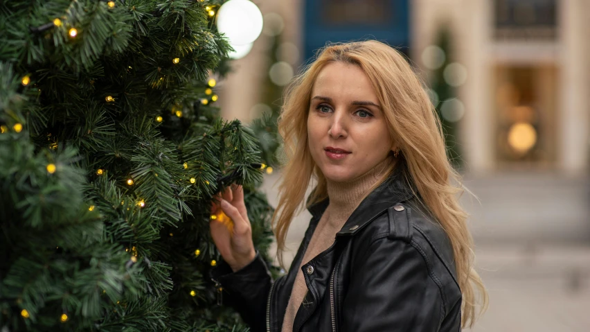 a woman standing next to a christmas tree, pexels contest winner, photorealism, wearing a leather jacket, avatar image, young blonde woman, candid portrait photo