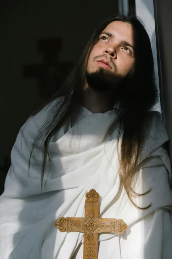 a man with long hair standing next to a cross, an album cover, unsplash, renaissance, golden-white robes, ( ( theatrical ) ), concerned, miro petrov