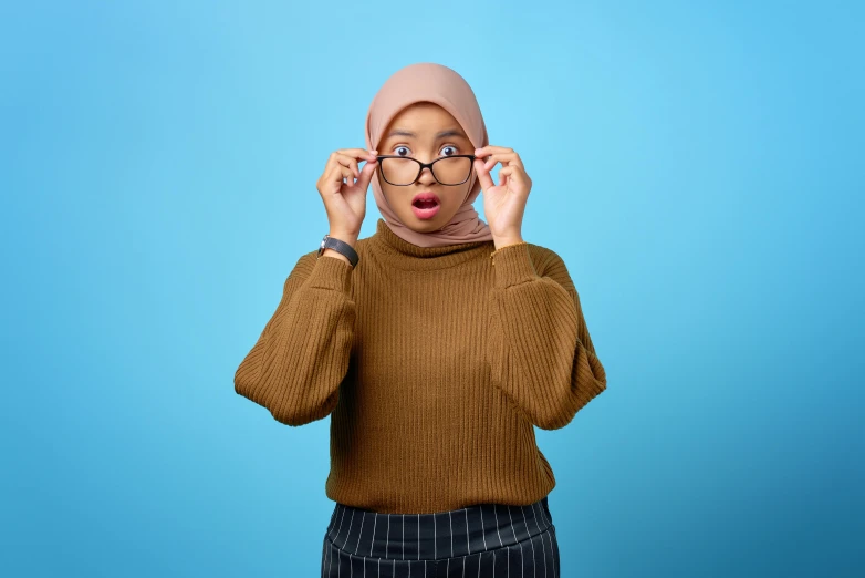 a woman wearing glasses with a surprised look on her face, trending on pexels, hurufiyya, islamic, he is wearing a brown sweater, with a blue background, student