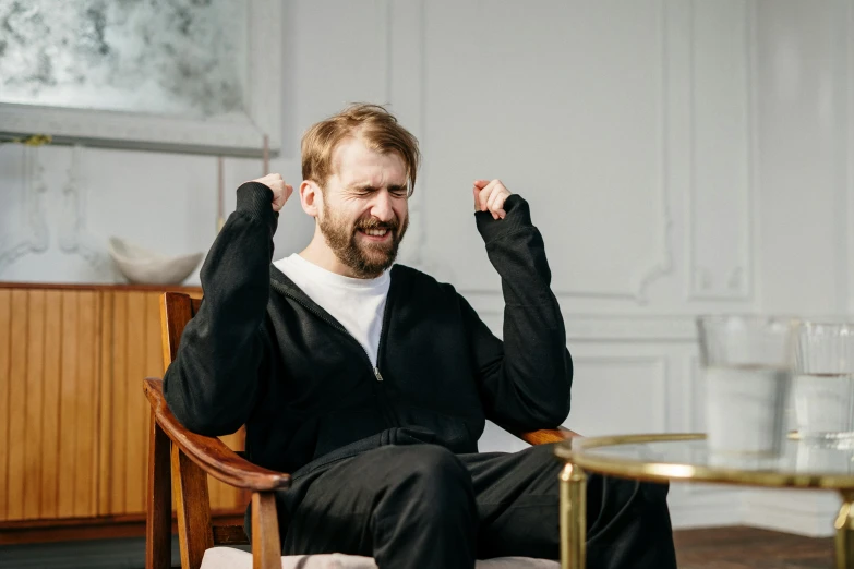a man sitting in a chair with his hands in the air, inspired by Constantin Hansen, pexels contest winner, happening, sarcastic smiling, man with beard, wearing a tracksuit, stressed out