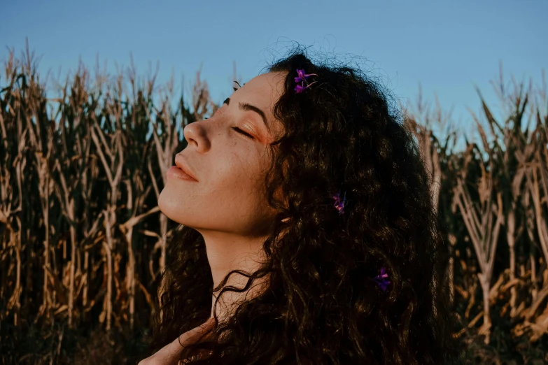 a woman standing in a corn field with her eyes closed, trending on pexels, brown curly hair, profile image, violet myers, with shiny skin