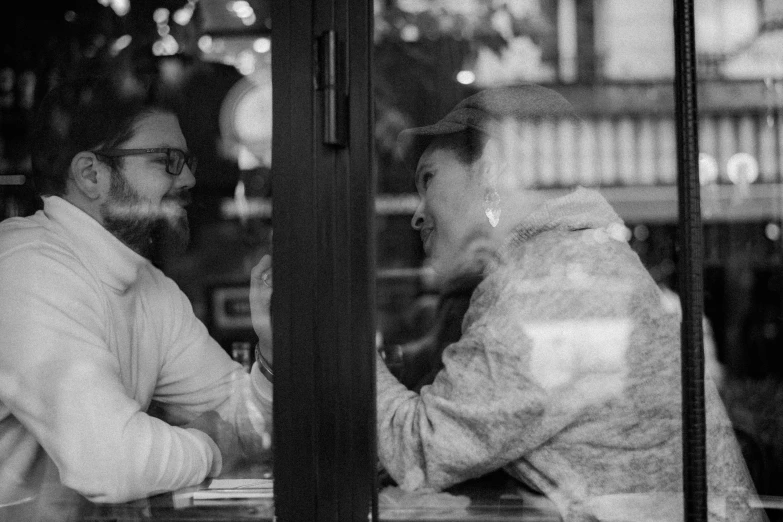 a couple of men standing next to each other in front of a window, a black and white photo, pexels, happening, sitting in a cafe, man and woman in love, reaching out to each other, laurie greasley and james jean