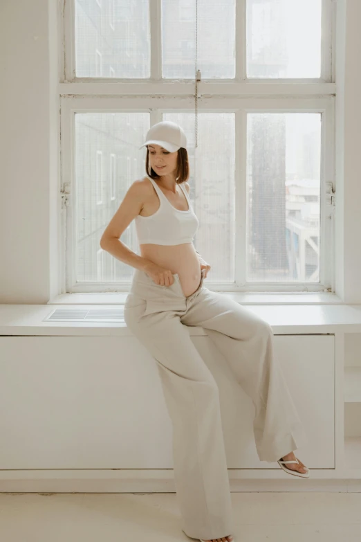 a pregnant woman sitting on a window sill, by Emma Andijewska, trending on pexels, art nouveau, white cap, baggy pants, on a white table, wearing a crop top