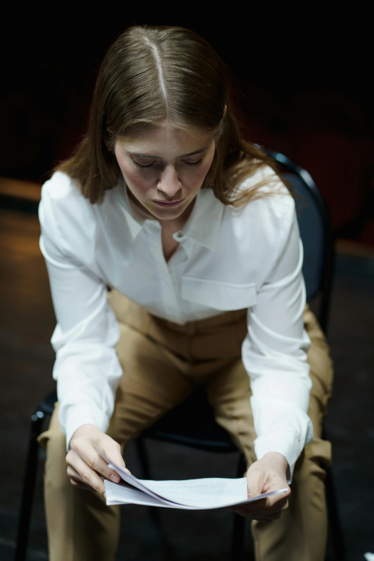 a woman sitting on a chair reading a piece of paper, somber expression, wearing a white button up shirt, ( ( theatrical ) ), bleeding audience