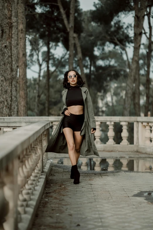 a woman walking on a bridge with trees in the background, pexels contest winner, baroque, trench coat, sexy look, black and green, bra and shorts streetwear