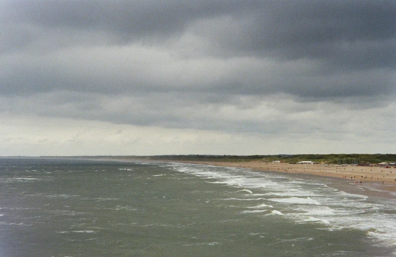 a group of people standing on top of a beach next to the ocean, dark clouds in the distance, omaha beach, panoramic view, high winds