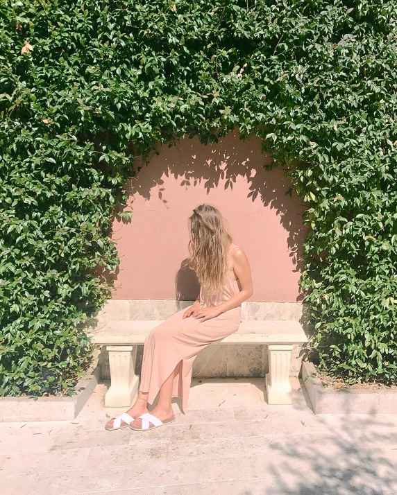 a woman sitting on a bench in front of a bush, a picture, inspired by Albert Joseph Moore, trending on unsplash, happening, pink marble building, slightly tanned, bare back, leaning against the wall