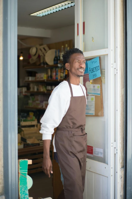 a man standing in the doorway of a store, wearing an apron, dark skinned, sustainability, brown:-2