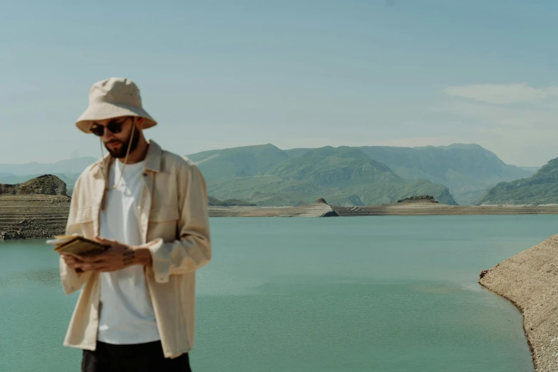 a man standing next to a body of water, pexels contest winner, looking at his phone, wearing off - white style, hills in the background, avatar image