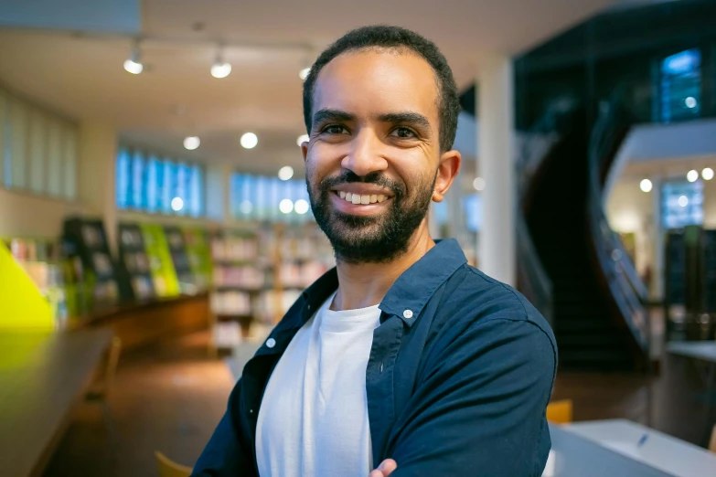 a man standing in a library with his arms crossed, a portrait, pexels contest winner, aida muluneh, headshot profile picture, smiling man, hicham habchi