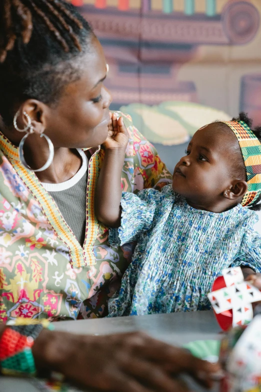 a woman sitting at a table with a baby, by Jessie Algie, trending on unsplash, afrofuturism, wearing an african dress, close together, multicoloured, still frame