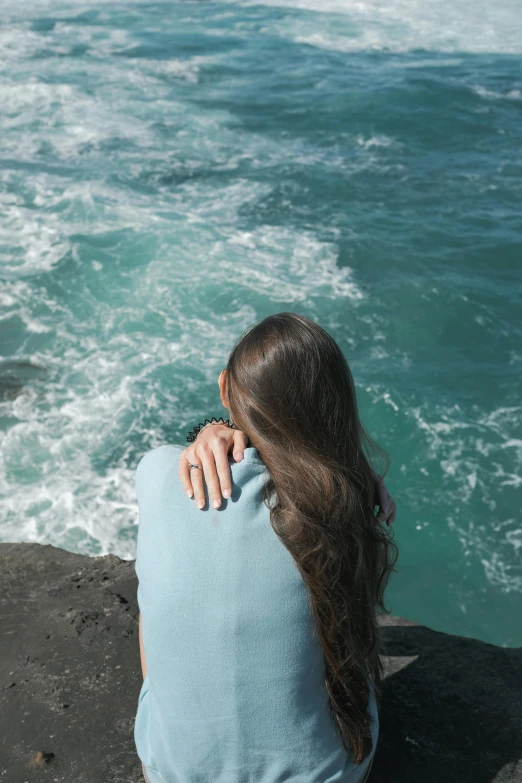 a woman sitting on top of a rock next to the ocean, wavy shoulder-length hair, with his back turned, wearing a light blue shirt, heartbreak