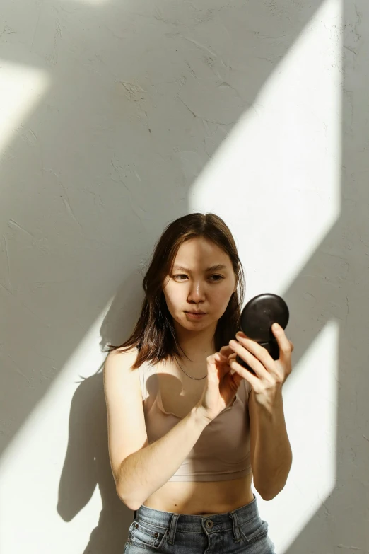 a woman standing next to a wall holding a cell phone, a picture, inspired by Kim Tschang Yeul, unsplash, minimalism, putting makeup on, sunlight filtering through skin, she is wearing a black tank top, round mirror on the wall