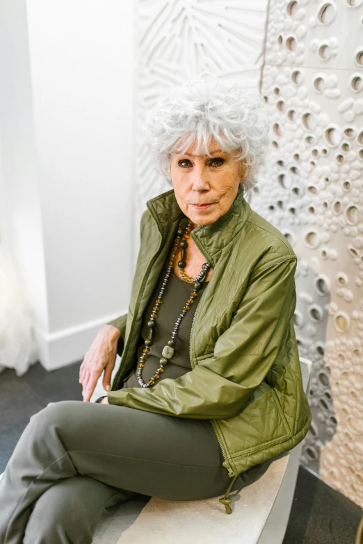 a woman sitting on a bench in a room, by Winona Nelson, wearing silver hair, el anatsui, wearing green jacket, in a white boho style studio