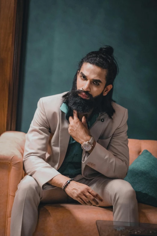 a man with a beard sitting on a couch, inspired by Sardar Sobha Singh, trending on pexels, shoulder long hair, topknot, wearing tight suit, androgynous male