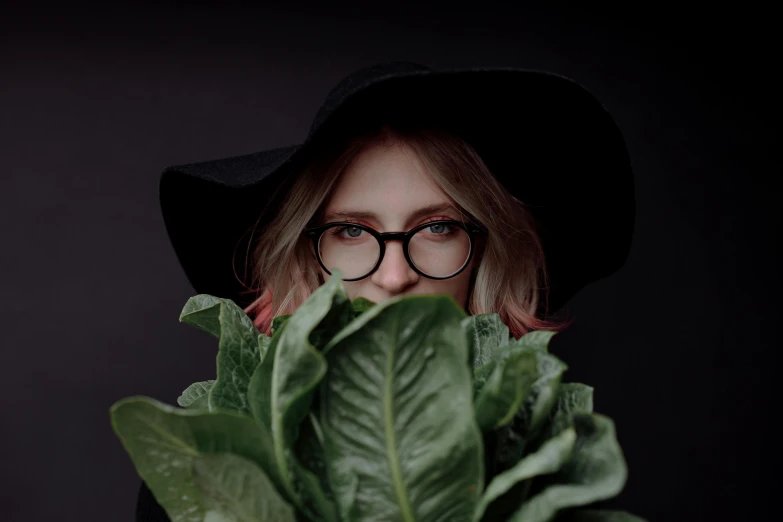 a woman holding a bunch of lettuce in front of her face, a picture, by Emma Andijewska, pexels contest winner, wearing black frame glasses, black hat, androgynous person, waist high