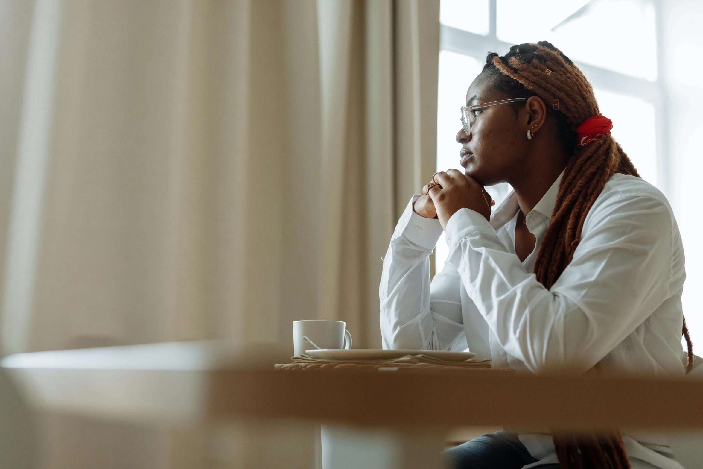a woman sitting at a table with a cup of coffee, looking out a window, with brown skin, praying, profile image