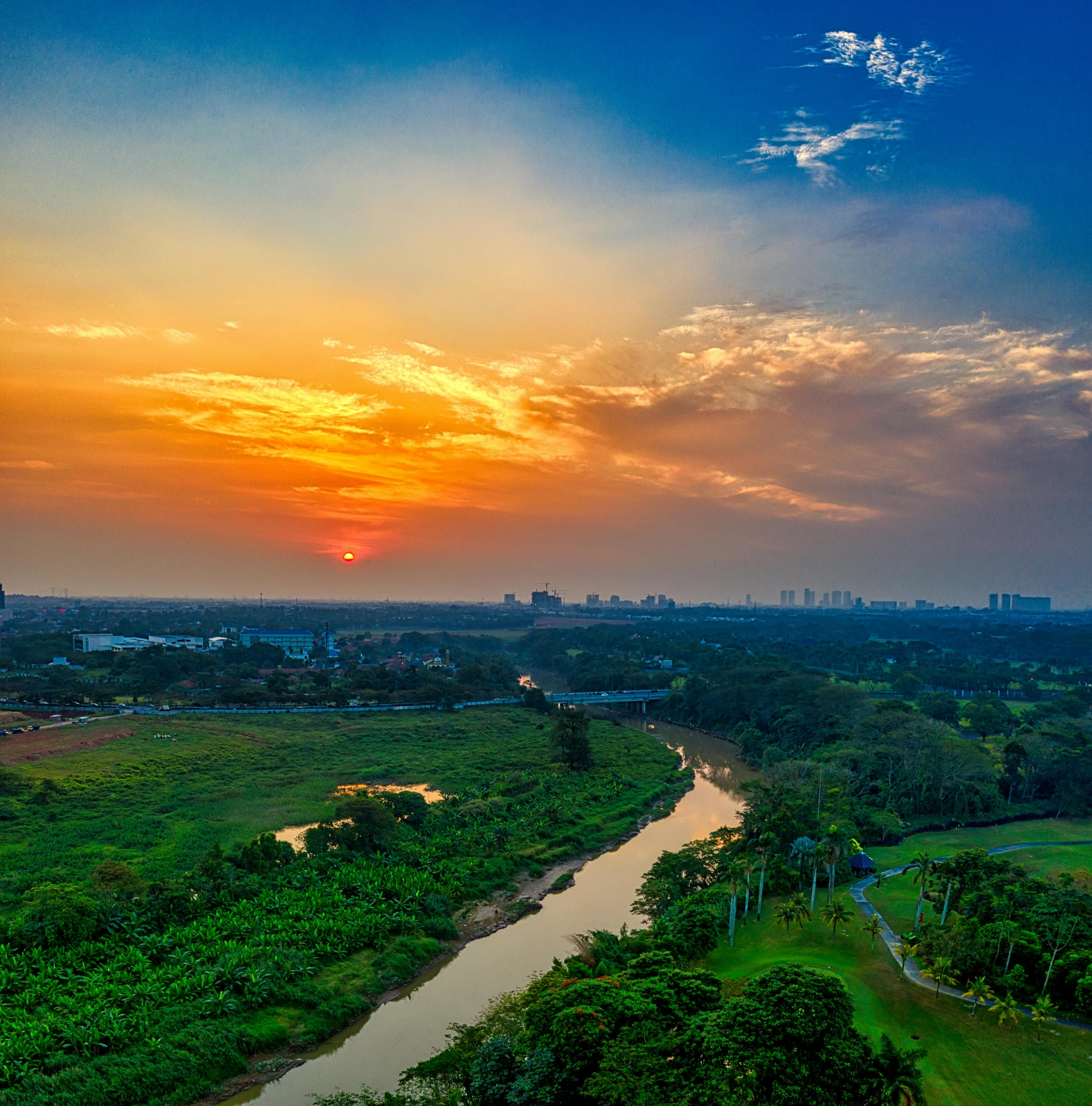 a river running through a lush green field, by Basuki Abdullah, pexels contest winner, hurufiyya, vista of a city at sunset, indian forest, orange and blue sky, colombo sri lanka cityscape