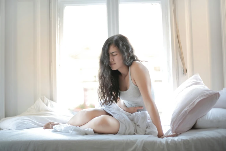 a woman sitting on top of a bed next to a window, trending on pexels, vomit, visible stomach, white, brunette