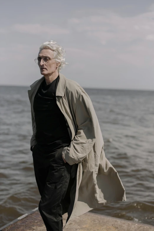 a man standing next to a body of water, an album cover, by David Begbie, flowy white grey hair, wearing a long coat, tilda swinton, on a beach