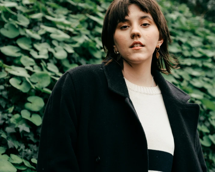 a woman standing in front of a hedge, an album cover, by Winona Nelson, trending on pexels, realism, wearing black overcoat, rebecca sugar, french bob, handsome girl