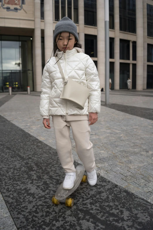 a young girl riding a skateboard down a sidewalk, an album cover, by Nina Hamnett, instagram, happening, cream and white color scheme, moncler jacket, baggy pants, little kid