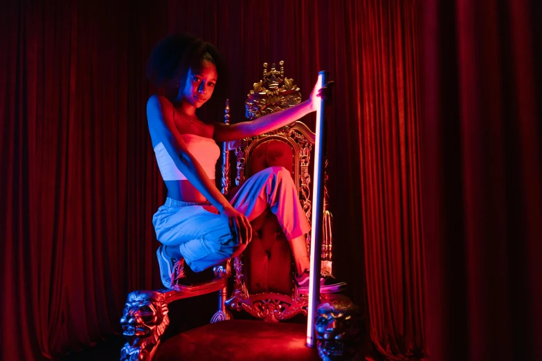 a woman sitting on top of a chair in front of a red curtain, inspired by Elsa Bleda, unsplash, baroque, night clubs and neons, african domme mistress, around the throne, pink and blue lighting