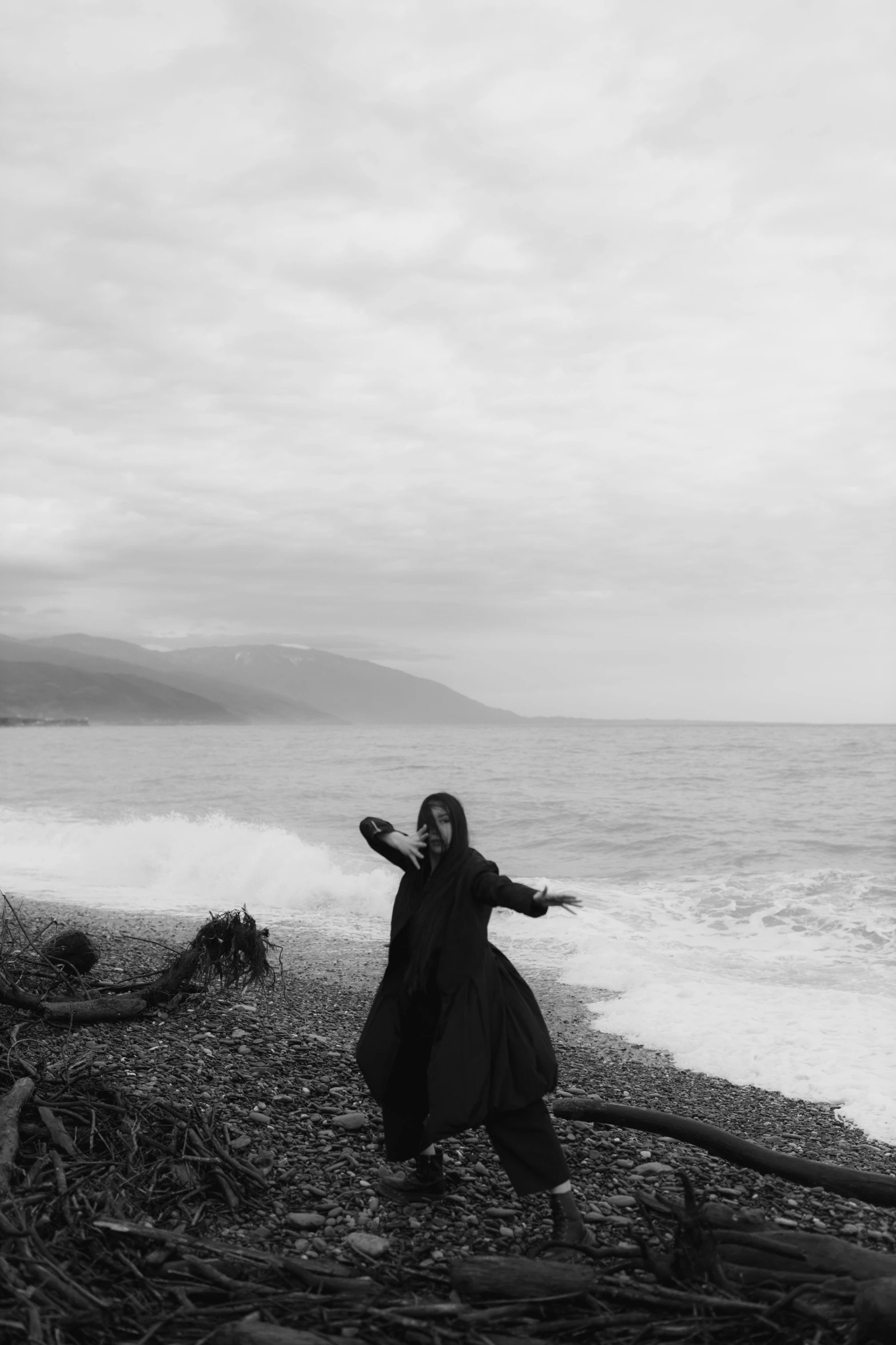 a woman standing on top of a beach next to the ocean, a black and white photo, severus snape, dark coat, waving at the camera, darkwave goth aesthetic