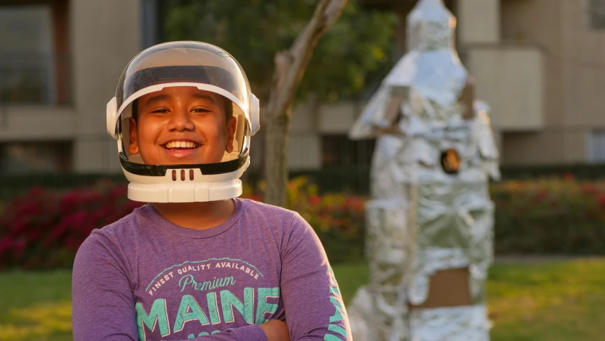 a young boy wearing a helmet standing in front of a statue, a portrait, inspired by Alan Bean, pexels contest winner, happening, made out of shiny white metal, cardboard cutout, subject is smiling, student