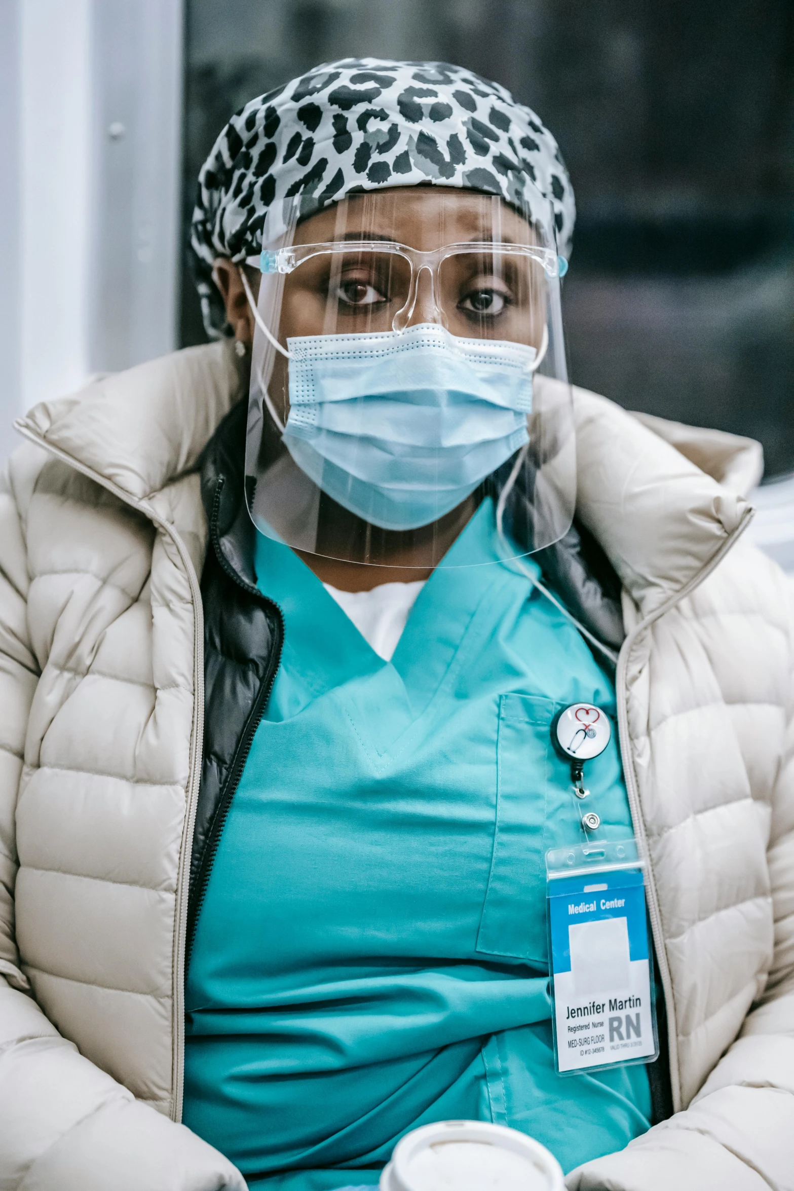 a woman wearing a face mask and holding a cup of coffee, surgical gown and scrubs on, african canadian, documentary photo, looking serious