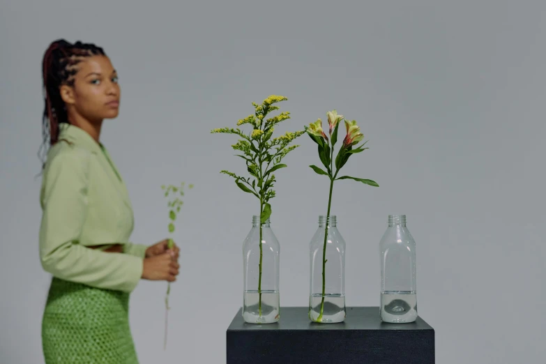 a woman standing next to three vases with flowers in them, inspired by Carrie Mae Weems, visual art, sustainable materials, realistic footage, promotional image, plants in scientific glassware