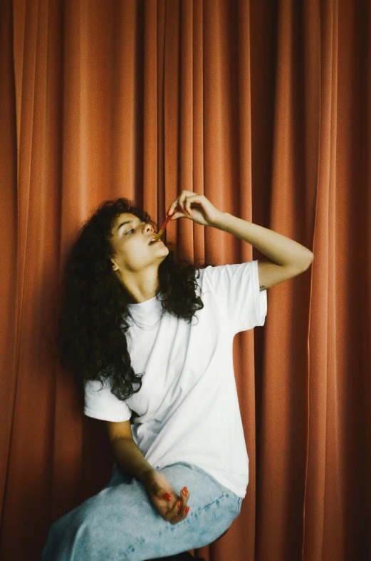 a woman sitting on a stool in front of a red curtain, an album cover, pexels contest winner, antipodeans, curly hair, dressed in a white t-shirt, profile pose, waving