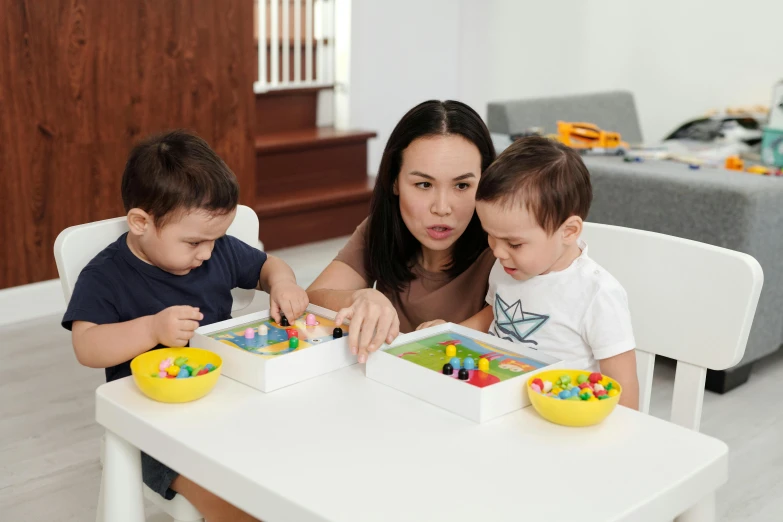 a woman sitting at a table with two children, inspired by Frederick Hammersley, pexels contest winner, interactive art, cubes on table, kids toys, profile image