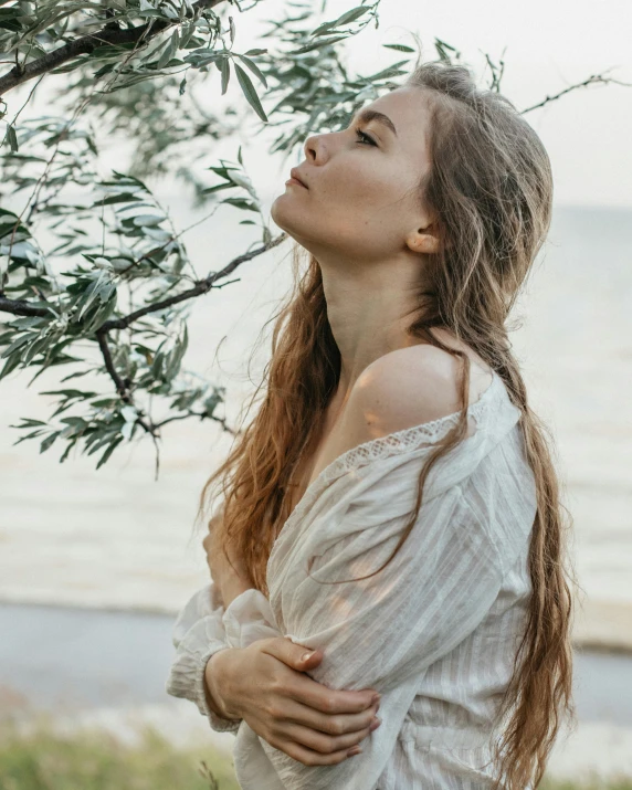 a woman standing under a tree next to a body of water, trending on unsplash, renaissance, long ashy hair | gentle lighting, queer woman, wearing a linen shirt, freckled pale skin