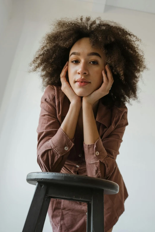 a woman sitting on a stool with her hands on her face, a portrait, by Lily Delissa Joseph, trending on unsplash, ashteroth, brown fluffy hair, on a white table, confident relaxed pose