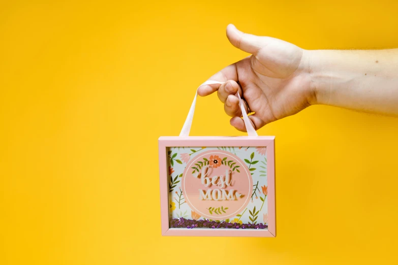 a woman's hand holding a pink gift box on a yellow background, a picture, by Julia Pishtar, folk art, square pictureframes, bright signage, botanical herbarium paper, feels good man