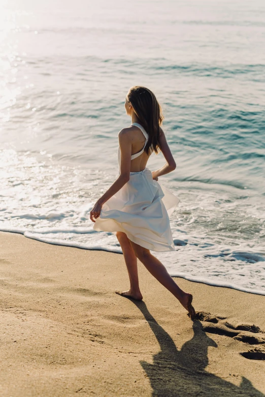 a woman in a white dress walking on a beach, overlooking the ocean, swirling around, sleek legs, sun - drenched