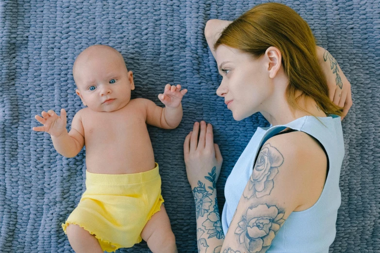 a woman holding a baby on top of a blue blanket, a tattoo, trending on pixabay, yellow carpeted, looking partly to the left, lying down, looking to the right