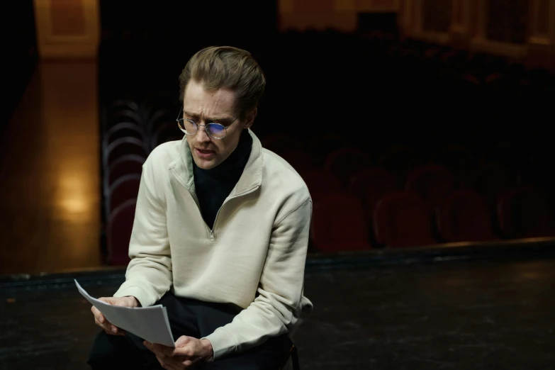 a man sitting on a chair reading a book, a portrait, by Celia Fiennes, unsplash, bauhaus, 3 actors on stage, young tom hiddleston, film still from movie dune-2021, man with glasses