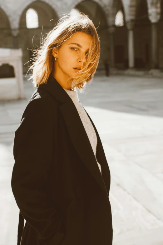 a woman in a black coat standing in a courtyard, trending on pexels, sleek blond hair, handsome girl, solid background, sun drenched