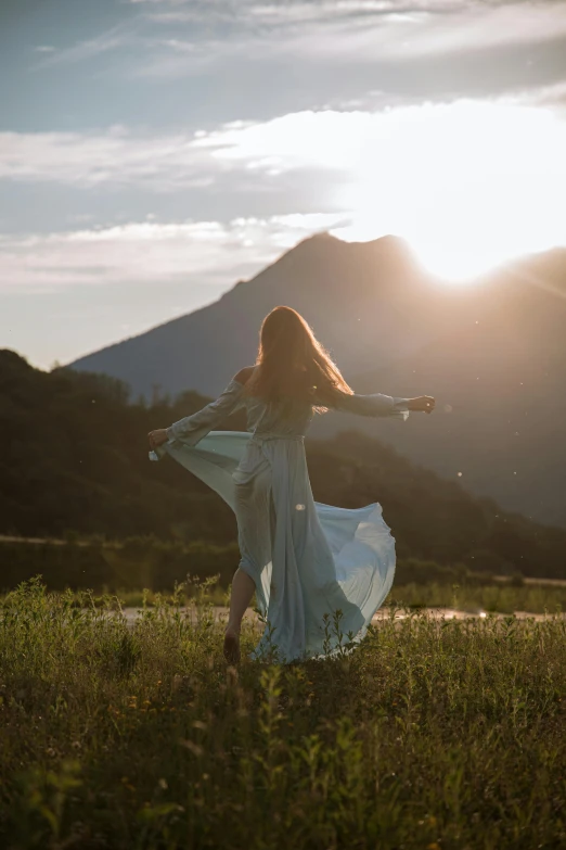 a woman standing on top of a lush green field, by Jessie Algie, unsplash contest winner, romanticism, she is dancing, sun rises between two mountains, modest flowing gown, switzerland