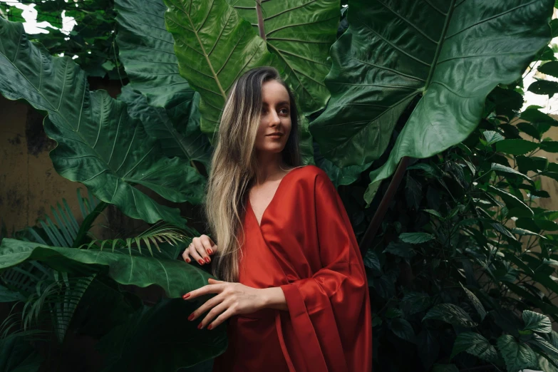 a woman in a red dress standing in front of large green leaves, by Emma Andijewska, pexels contest winner, wearing a luxurious silk robe, long hair and red shirt, wearing a long flowy fabric, ignant