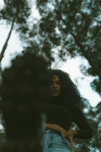 a woman standing in the middle of a forest, an album cover, pexels contest winner, black curly hair, low quality footage, cinematic outfit photo, with a black background