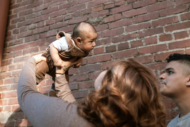 a man holding a baby up in the air, pexels contest winner, photorealism, brown, urban surroundings, vfx shot, gaze down