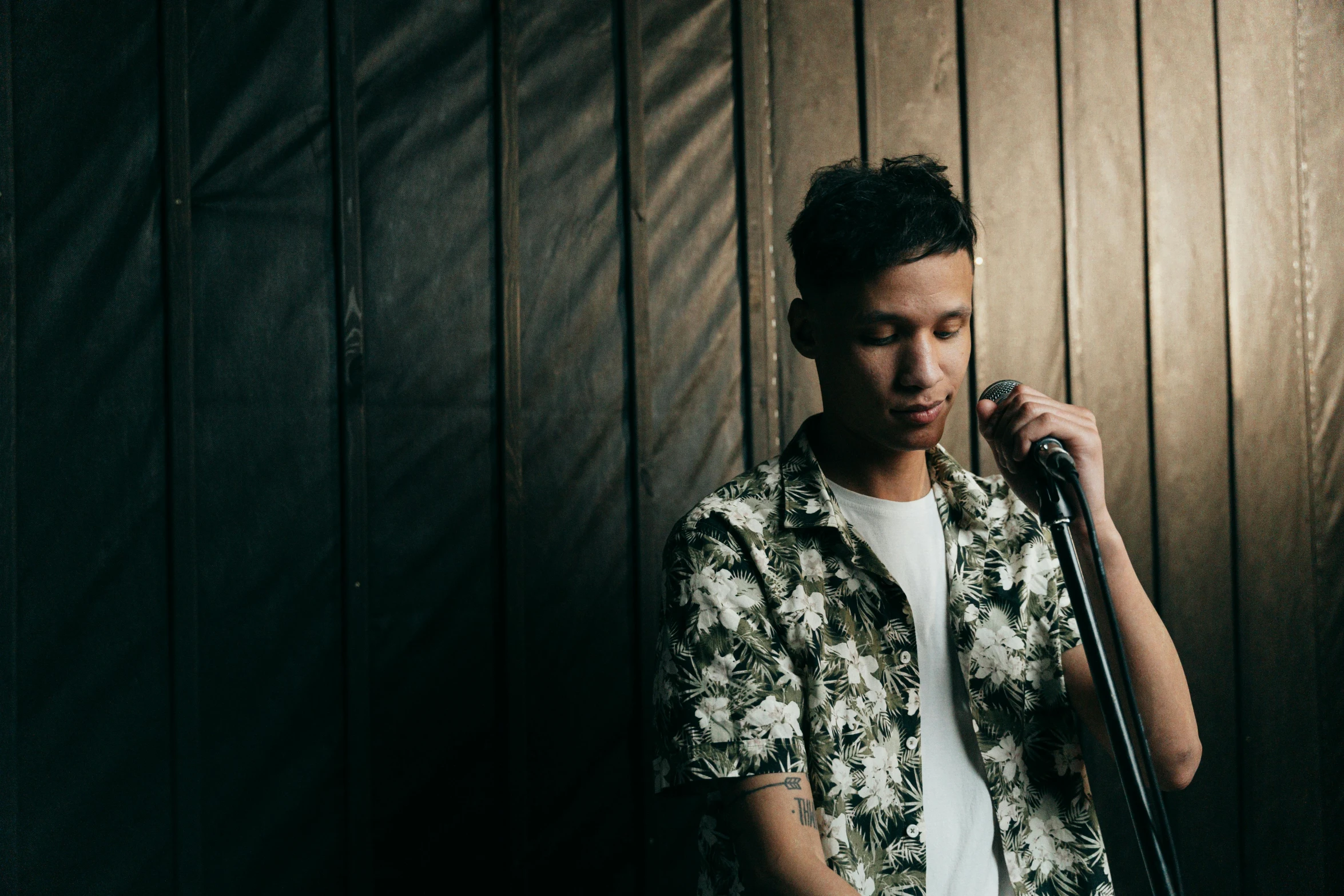 a man holding a microphone in front of a wooden wall, an album cover, by Robbie Trevino, unsplash, realism, asian man, background image, 18 years old, standing in a dimly lit room