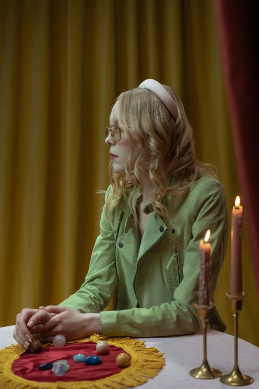 a woman sitting at a table with a candle, inspired by Nan Goldin, renaissance, wearing green suit, elle fanning, wearing gold glasses, pale skin curly blond hair