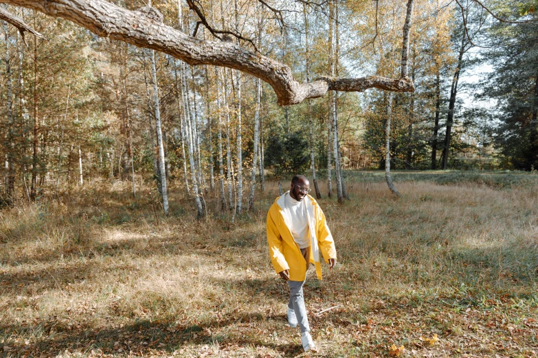 a woman in a yellow jacket walking through a forest, pexels contest winner, adut akech, grassy autumn park outdoor, man standing, offwhite