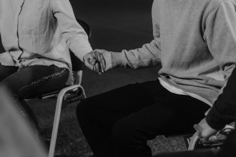 a black and white photo of two people holding hands, siting in a chair, roleplay, late evening, wearing jeans and a black hoodie