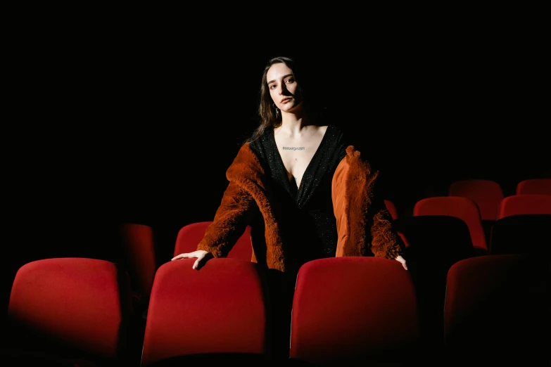 a woman standing in front of a row of red chairs, an album cover, inspired by Elsa Bleda, pexels contest winner, antipodeans, sitting in a movie theater, dilraba dilmurat, wearing a fancy jacket, lily frank