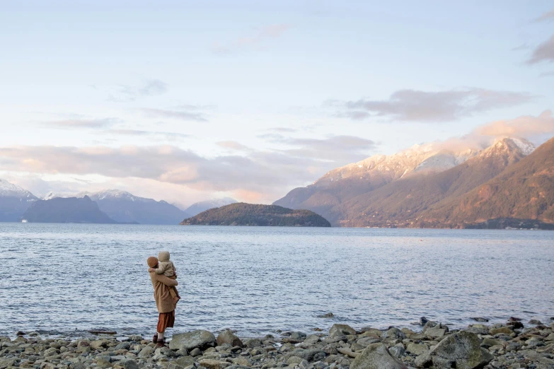 a person standing on a rocky beach next to a body of water, a picture, mountains and oceans, vancouver, tiny person watching, brown